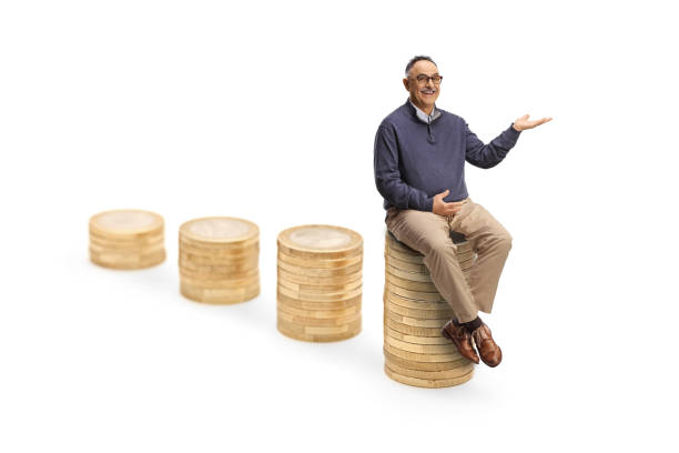 Mature man sitting on the tallest pile of coins and showing with hand isolated on white background